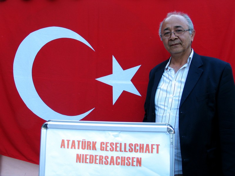 17h7e Bülent Demiral, Vorsitzender Europäische Atatürk Bildungs- und Kulturgesellschaft, hinter einem Plakat der Atatürk Gesellschaft Niedersachsen, türkische Fahne