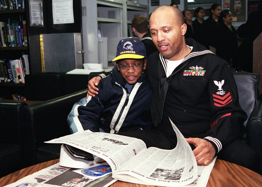 US Navy 030201-N-8770A-001 Information Systems Technician 1st Class Russell Thompson reads a Stars ^ Stripes newspaper to his son in the ship's library during a Family ^ Friendship Cruise