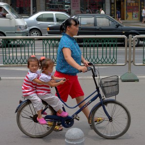 A woman is pushing a bike with two kids on it.