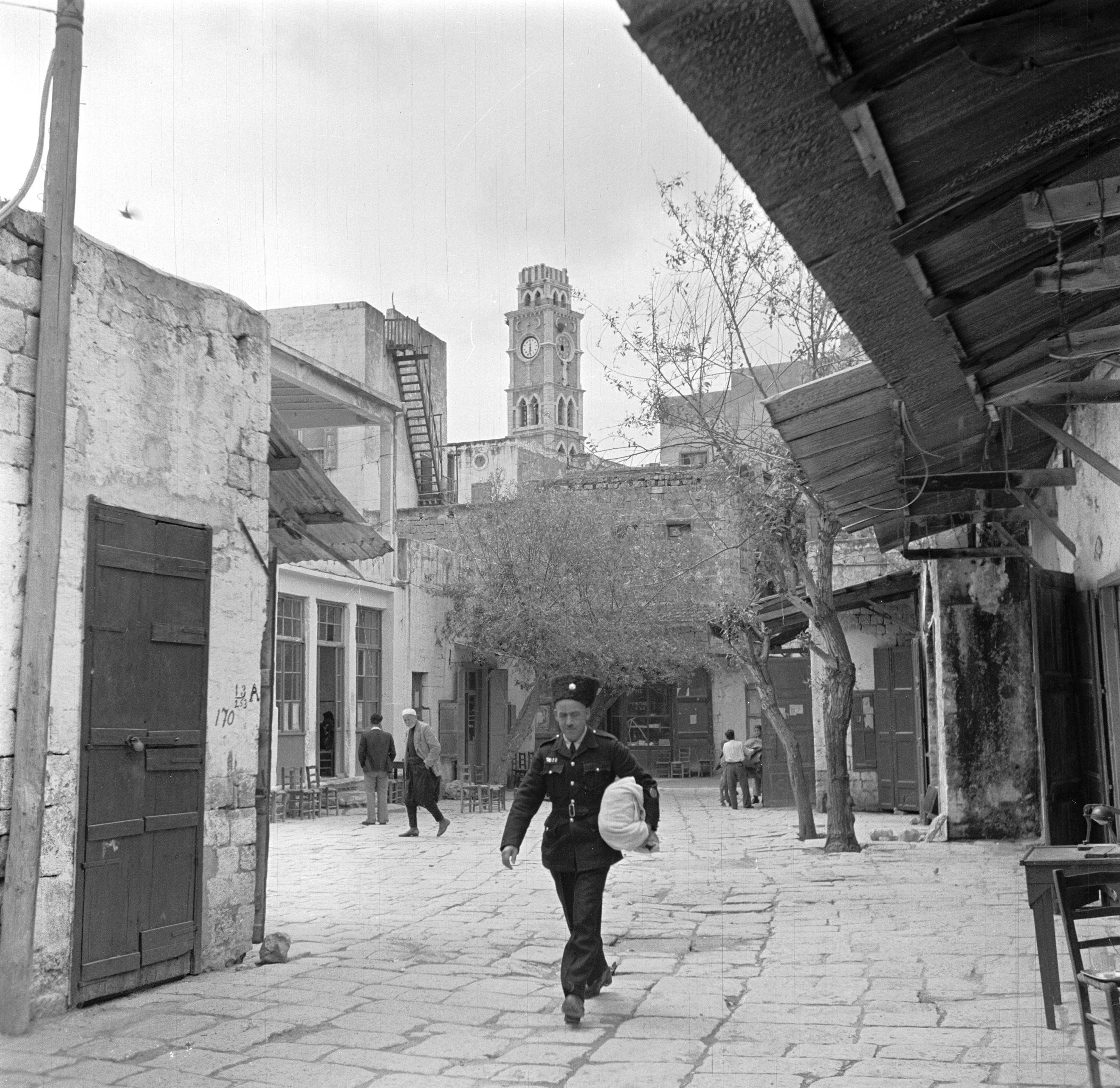 Acre. Publiek, waaronder een politieagent in een ouderwets uniform, op pleintje in de oude stad. Op de achtergrond een klokkentoren. Israël 1948-1949, Acre (Akko) (255-0040)