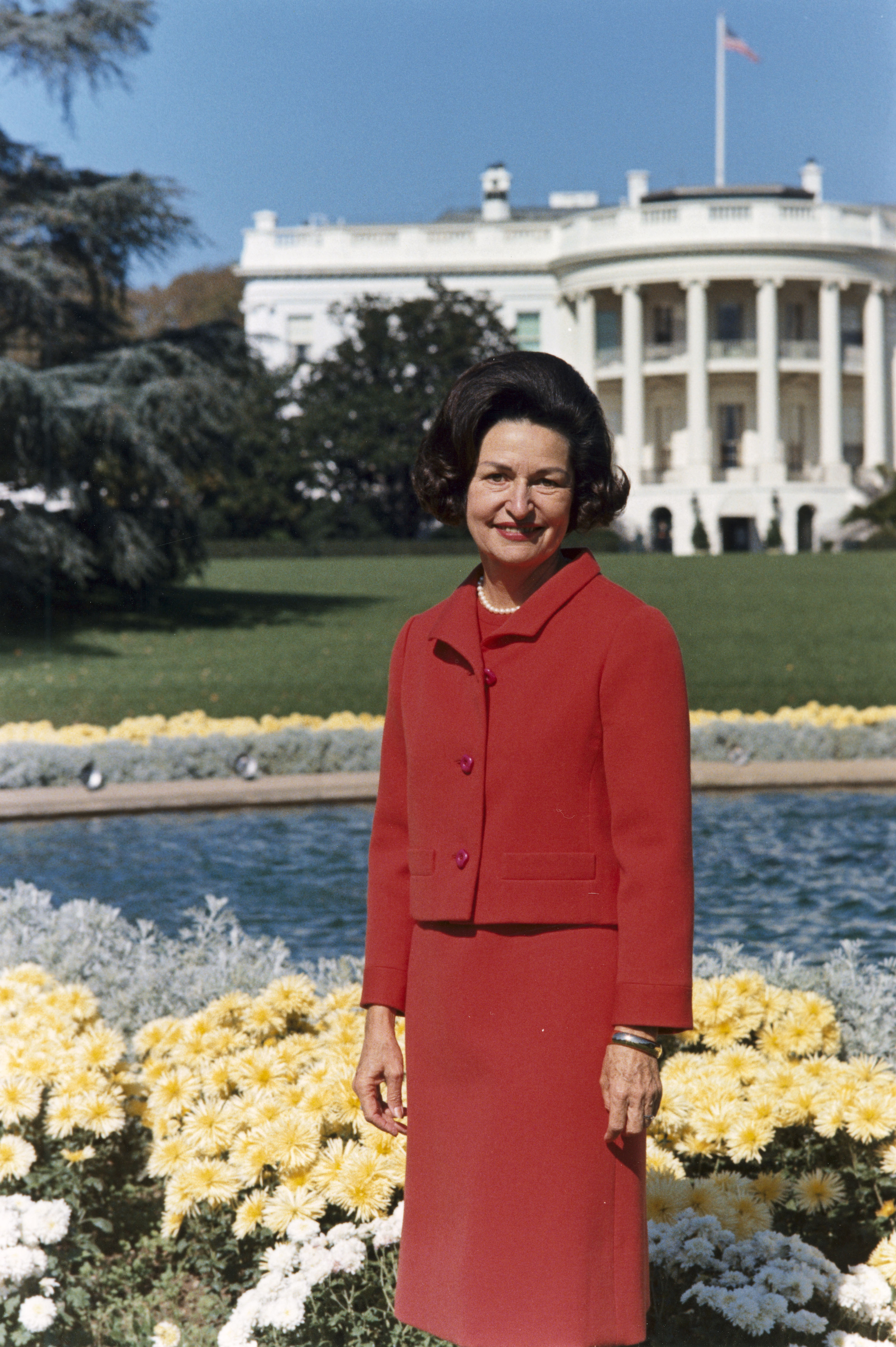 Lady Bird Johnson, photo portrait, standing at rear of White House, color